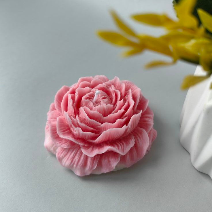 Pink flower-shaped object on a light gray background with a blurred yellow plant in the corner.