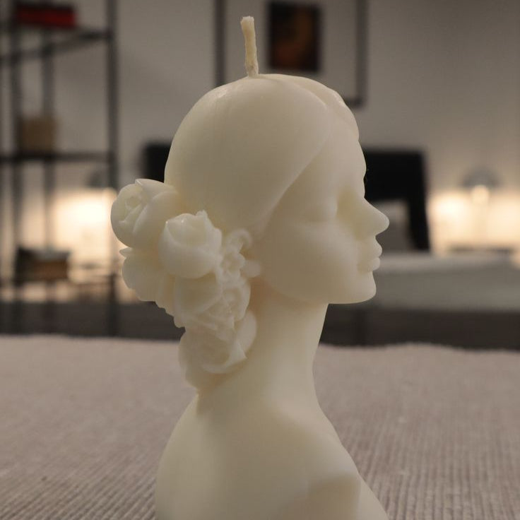 Candle shaped like a woman's head with flowers in her hair on a textured surface.